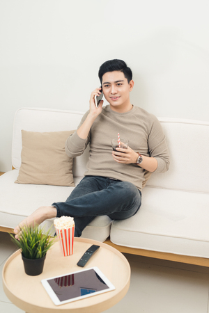 Handsome asian man is talking on the phone and smiling sitting on couchの写真素材