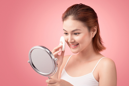Young lady applying blusher on her face with powder puff and mirror, skin care concept on pink background.の写真素材