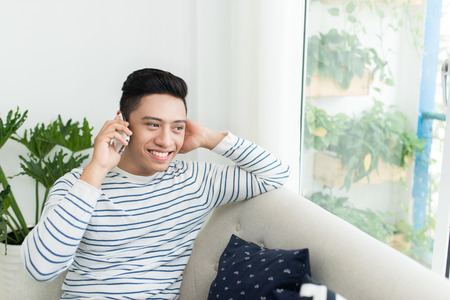 Young handsome asian man using modern mobile smart phone sitting on a couch at homeの写真素材