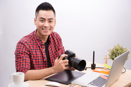 Professional photographer. Portrait of confident young man in shirt holding hand on camera while sitting at his desk.の写真素材