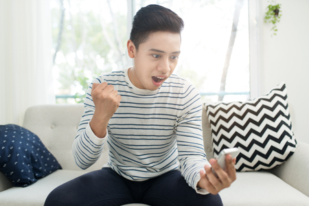 Portrait of excited asian man looking game on mobile phone at homeの写真素材