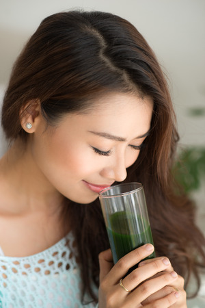 Smiling young asian woman drinking green fresh vegetable juice or smoothie from glass at homeの写真素材