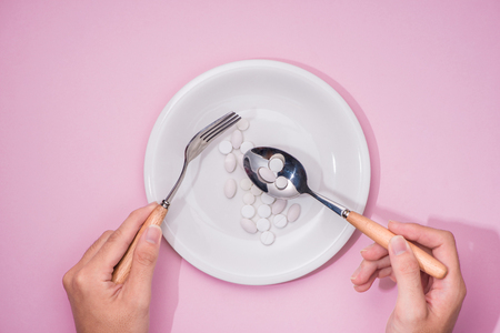 Top view of man's hands at dining table holding a fork and knife above dish with pills over pink background.の写真素材