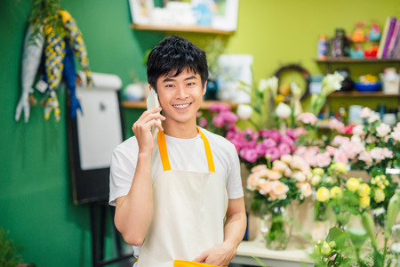 Smiling asian male florist calling on smartphone at flower shop.の写真素材