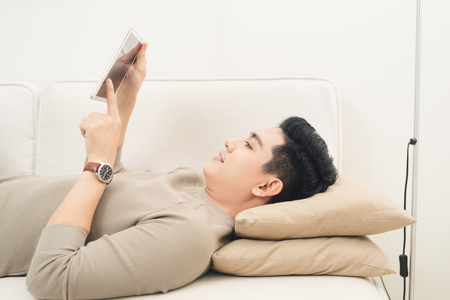Enjoying time at home. Cheerful young man holding digital tablet and looking relaxed while lying on the couch at homeの写真素材