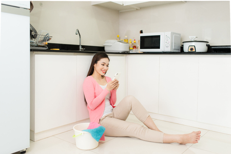Beautiful young woman typing on mobile, sitting on the floor at home near a bucket with things for cleaningの写真素材