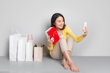 Young asian woman sitting besides row of shopping bags holding red gift box taking sellfieの写真素材