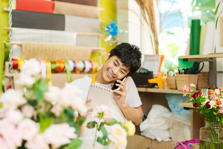 Smiling asian male florist calling on smartphone and making notes at flower shop counterの写真素材