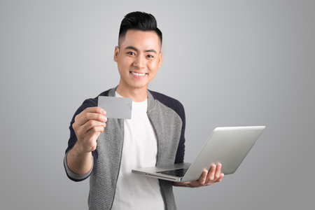 Young asian man with laptop and credit card in studio isolated gray backgroundの写真素材