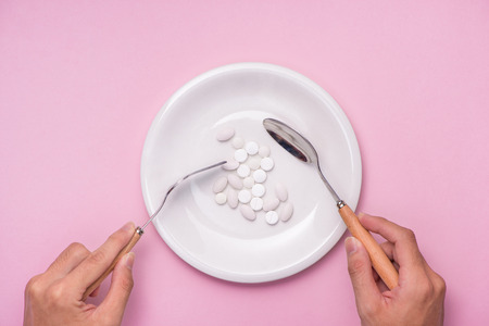Top view of man's hands at dining table holding a fork and knife above dish with pills over pink background.の写真素材