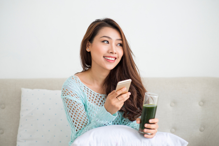 Smiling young asian woman drinking green fresh vegetable juice or smoothie from glass using smartphone at homeの写真素材
