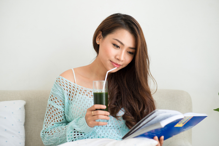 Smiling young asian woman drinking green fresh vegetable juice or smoothie from glass at homeの写真素材