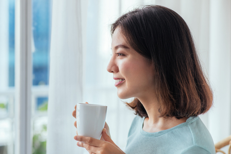Young pretty woman sitting at opened window drinking coffee and looking outside enjoys of restの写真素材