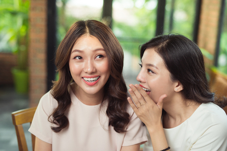 Young asian woman telling secrecy news to her surprised girlfriendの写真素材