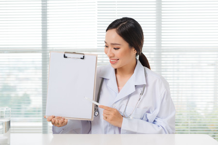 Portrait of asian female doctor pointing on clipboard while sitting at consulting roomの写真素材