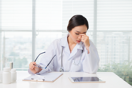 Frustrated young woman doctor keeping eyes closed with coat wearing stethoscope at worktableの写真素材