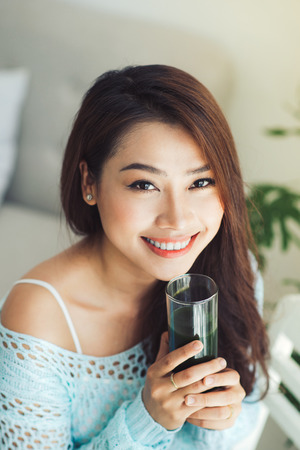 Pretty young asian woman holding green fresh vegetable juice or smoothie from glass at homeの写真素材
