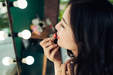 Beautiful young woman applying glossy lipstick on lips in front of a make up mirror at homeの写真素材