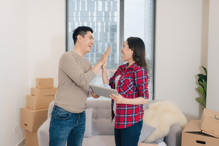 Concept young couple moving house. Beautiful young couple giving high five and smiling while standing among moving cardboard boxesの写真素材