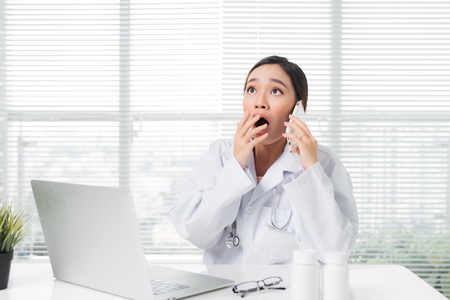 Young surprised female doctor sitting at office desk and working with a laptopの写真素材