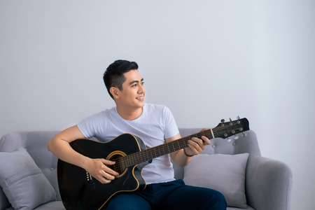 Handsome asian man playing on the guitar on the couch at homeの写真素材