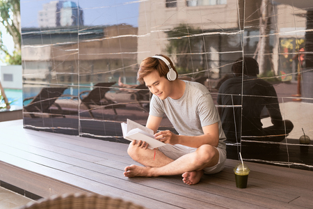 Young asian man reading book and listening to music by the pool on a sunny summer dayの写真素材