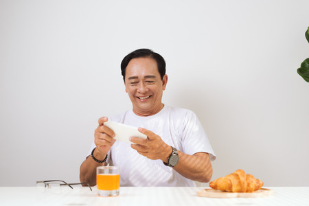 Happy senior couple having croissant breakfast at home and drinking orange juice, using smartphone.の写真素材