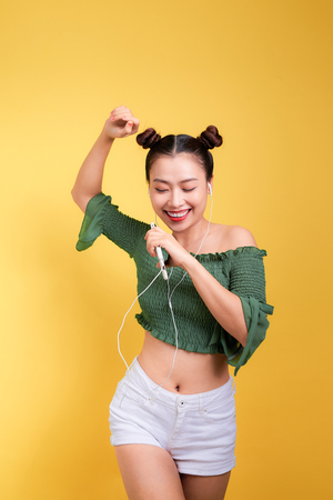 Fashion smiling asian woman listening to music in earphones  over yellow backgroundの写真素材