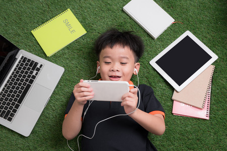 Cute little boy listens to music on the grass with his laptop, tablet and notebooks around.の写真素材