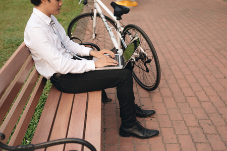 Young handsome businessman with laptop and bicycle sitting on bench in parkの写真素材