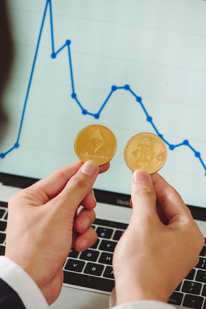 Man holding physical bitcoin and ethereum cryptocurrency coins in his hands on the background of the chartの写真素材