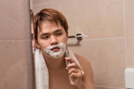 Handsome young asian man is shaving his face and looking at the mirror.の写真素材