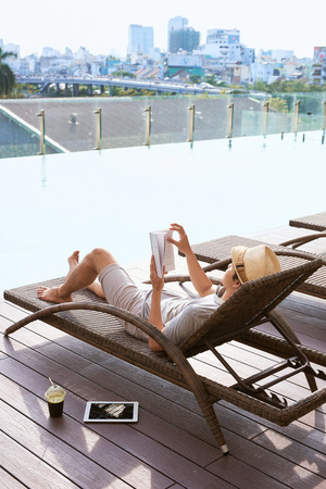 Young asian man reading book by the pool on a sunny summer day