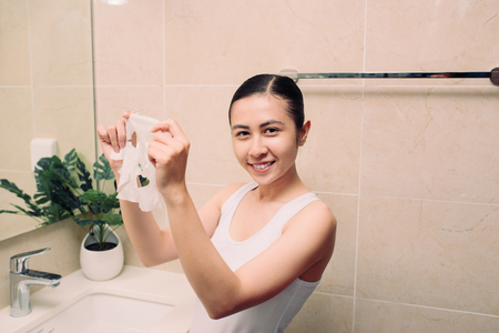 Beauty Skin Care Concept - Beautiful Asian Woman applying paper sheet mask on her face in bathroomの写真素材