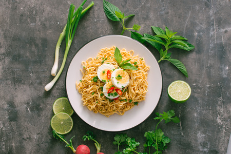 Dry instant noodles put egg with fresh herbs, garnish of cilantro and Asian basil, lemon, lime on dark stone backgroundの写真素材