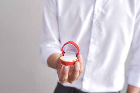 Handsome elegant man in a white shirt is holding a box with an engagement ring and shows in camera.の写真素材