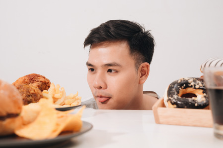 Young man having cravings for donuts, hamburger, chicken with fries insteadの写真素材