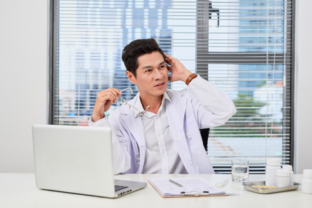 Asian male doctor typing on laptop computer while sitting at the table near the window in hospital officeの写真素材