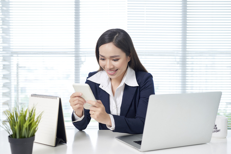 Asian businesswoman sitting by desk with laptop and using mobile in officeの写真素材