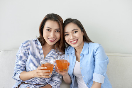 Two sisters on the sofa with orange juice in hands. Two best friends enjoying time.の写真素材