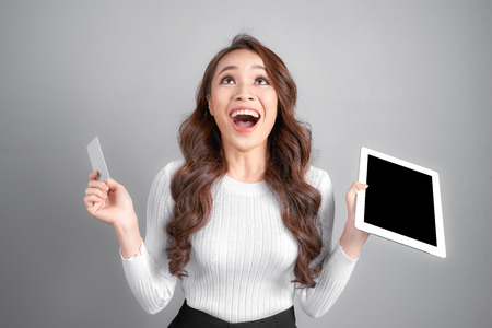 Smiling woman showing blank credit card hold tablet pc in hand, isolated over gray backgroundの写真素材