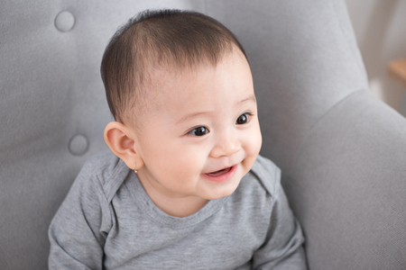 Closeup portrait of cute adorable smiling laughing, baby girl with black eyes sitting on sofa looking away from camera, natural window light, lifestyleの写真素材