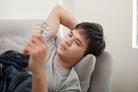 A young man is sick with flu, lies at home under a blanket, takes a pillの写真素材