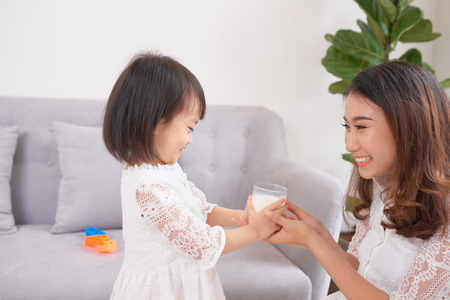 Little girl and her mom drinking milk sitting on sofa at home. Motherhood and care, healthy eating and lifestyle, early development concept, copy spaceの写真素材