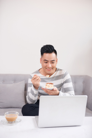 Man eating cake while sitting on sofa at home with notebook on luxurious sofa in living groom.の写真素材