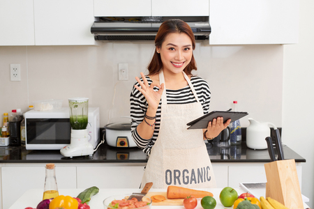 Beautiful young woman is using a digital tablet and smiling while cooking in kitchen at homeの写真素材