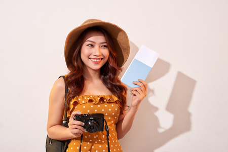 Portrait of a happy young woman in hat holding camera and showing passport while standing isolated over beige backgroundの写真素材