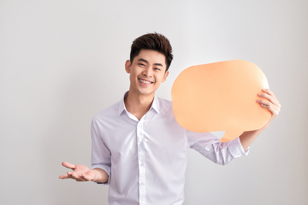 Handsome asian man in classic shirt is holding a speech bubble and smiling, standing on white wallの写真素材