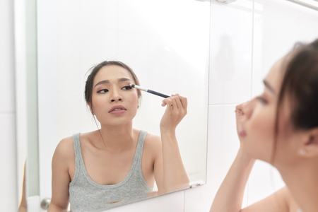 A young woman is standing in front of the bathroom mirror and putting on makeup.の写真素材
