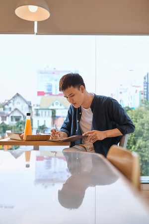 Handsome young designer writes creative graphic notes and making sketch in notebook while sitting at wooden table in coffee interior. Thoughtful man writing new successful ideas for blog articleの写真素材